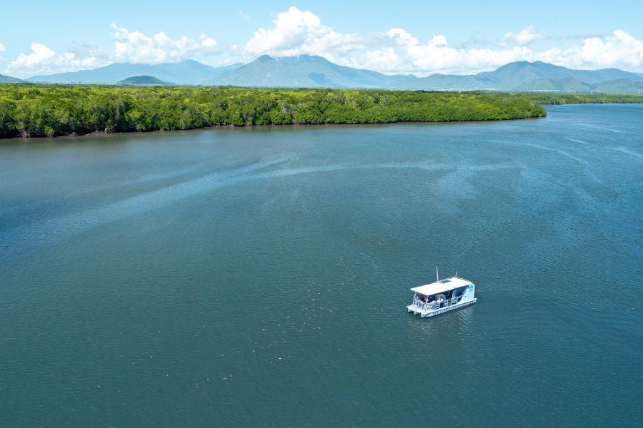 Experience a scenic Cairns City Sights River Cruise as a small white boat glides along a wide river, framed by lush greenery and mountains.