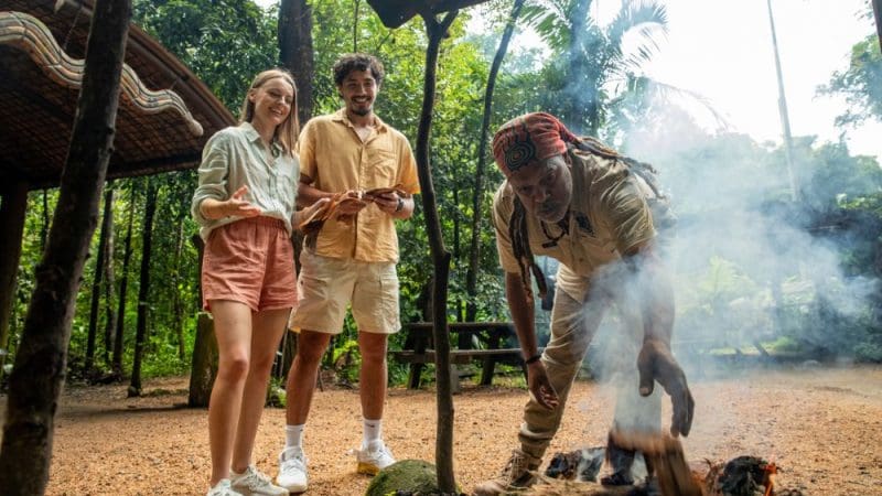 Three enjoying the outdoors; two smiling and watching as one tends a smoky campfire during a Private Exclusive Daintree Mossman Tour.
