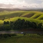 Sleek white car on a Private Exclusive Atherton Tablelands Tour cruises lush green hills beneath a dramatic sunrise or sunset sky.