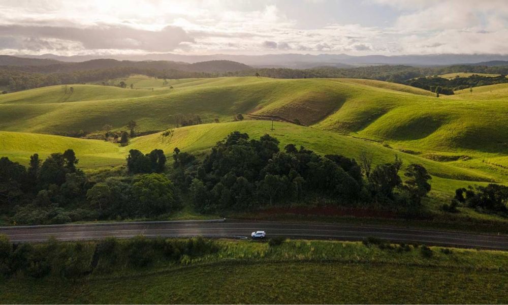 Sleek white car on a Private Exclusive Atherton Tablelands Tour cruises lush green hills beneath a dramatic sunrise or sunset sky.