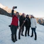 Four friends in winter gear snap a selfie atop a snowy glacier, majestic mountain peaks and stunning alpine scenery in the background.