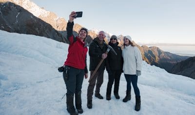 Four friends in winter gear snap a selfie atop a snowy glacier, majestic mountain peaks and stunning alpine scenery in the background.