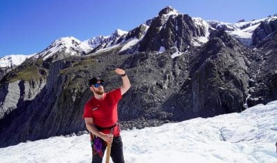 Man in red shirt stands on snowy glacier, smiling amid majestic mountain peaks and vibrant blue sky, enjoying an epic adventure.