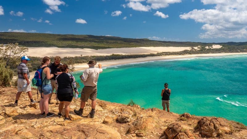 Travellers on the 8 Day Sydney to Brisbane with Fraser Island Comfort Tour admire stunning turquoise waters and lush forest from a scenic cliff.