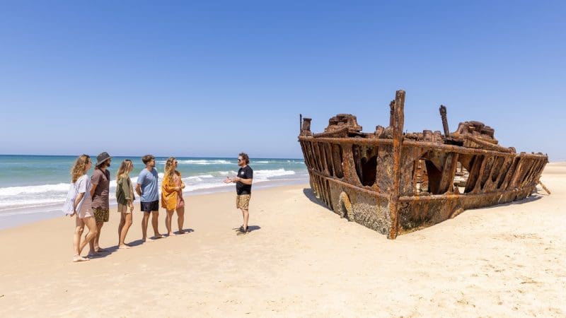 Travellers relax on a pristine Fraser Island beach by a historic shipwreck during the 8 Day Sydney to Brisbane Comfort Tour adventure.