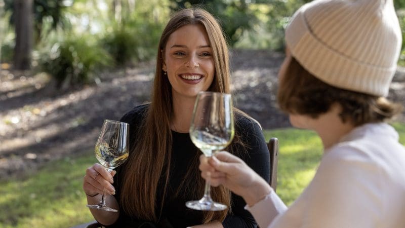 Two women smiling and toasting glasses of wine in a lush park during their 16 Day East Coast Australia Comfort Tour from Sydney.
