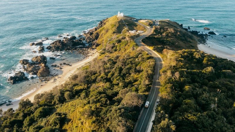 Scenic winding road curves towards a striking white lighthouse, evoking the 8 Day Sydney to Brisbane Fraser Island Comfort Tour experience.