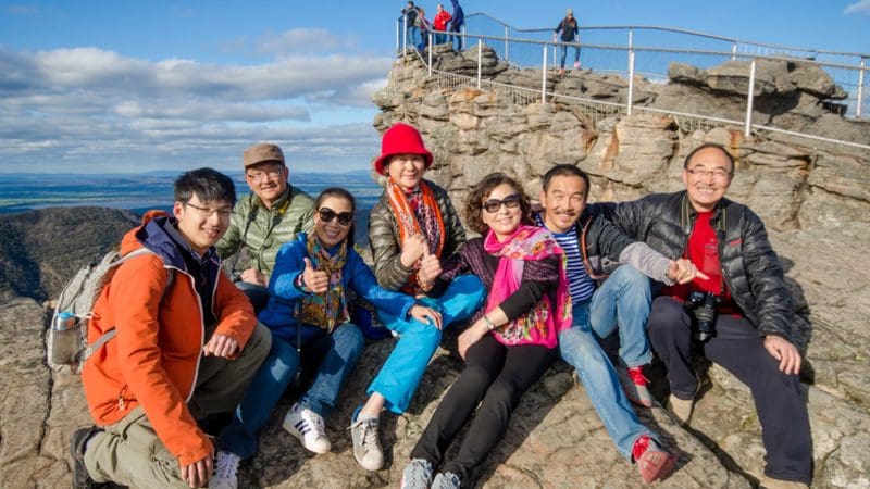 A group of seven happy people on rocky terrain enjoying a 1 Day Grampians Eco Wilderness Escape with stunning scenic views behind them.