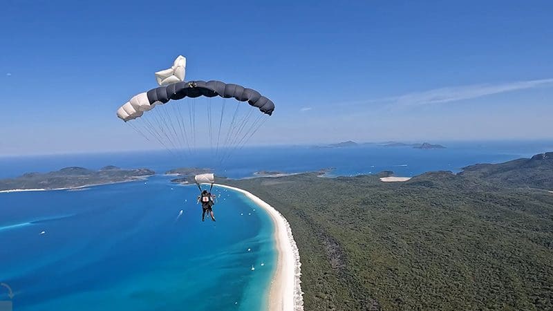 Experience a thrilling tandem skydive with video and photos, as a parachute soars over Whitehaven Beach’s turquoise water and lush hills.