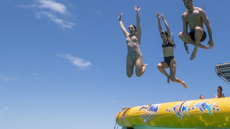 Three friends in vibrant swimwear leap from a colourful inflatable float into the crystal-clear Great Barrier Reef sea on an eco adventure.