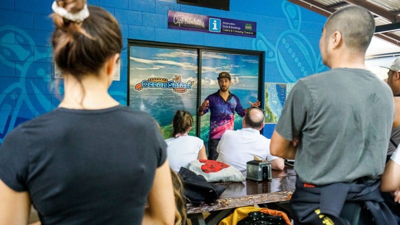 An expert guide presents the Great Barrier Reef Eco Adventure to an attentive group seated indoors by a vibrant ocean-themed blue wall.