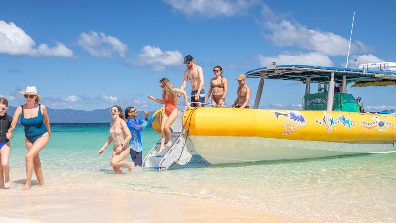 Tourists in colourful swimsuits disembark a yellow boat onto a pristine tropical Great Barrier Reef beach for an exciting eco adventure.