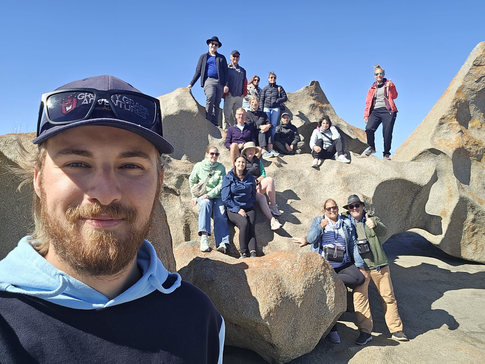 A smiling group on rugged rocks beneath a clear sky captures a selfie during their Kangaroo Island 2 Day Wildlife Adventure tour.