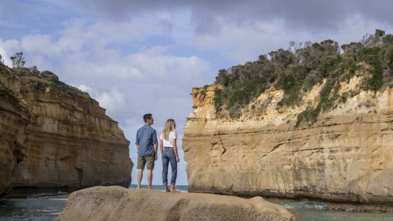 A couple atop a scenic rock enjoys panoramic Great Ocean Road cliffs and ocean views on a 1 Day Reverse Eco Tour adventure.