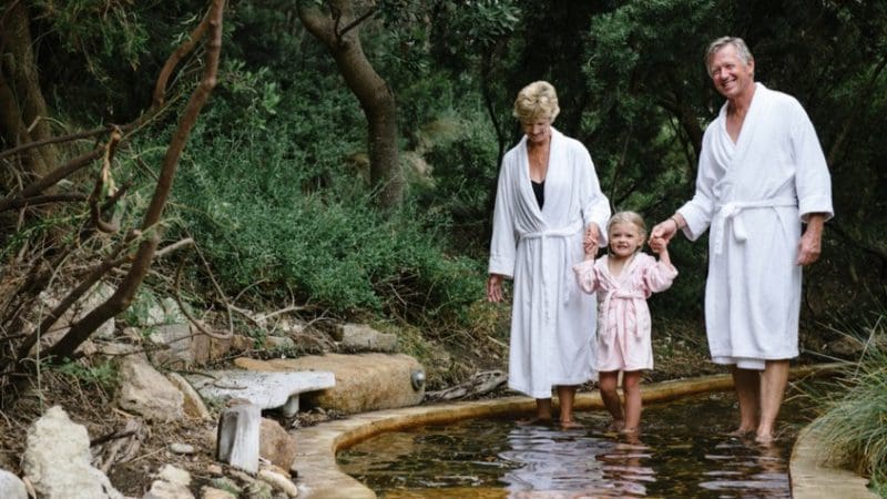 Happy family of two adults and a child in spa robes walking barefoot by lush greenery after Peninsula Hot Springs day spa shuttle experience.