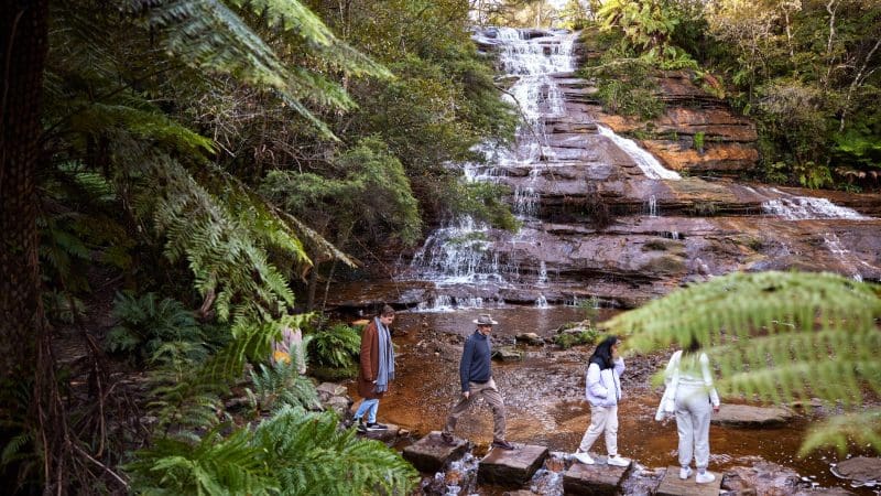 Four visitors cross stepping stones beside a cascading waterfall on the popular 1 Day Blue Mountains Sunset Wilderness Tour in Australia.