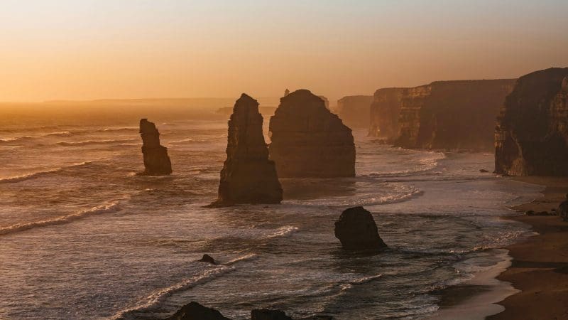 Dramatic rock formations emerge from the sea at sunset on a Great Ocean Road tour from Melbourne, with waves crashing ashore.