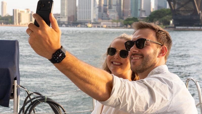 Smiling couple in sunglasses captures a selfie whilst enjoying a Sydney Harbour BYO Sunset Sparkle Catamaran Cruise at sunset.