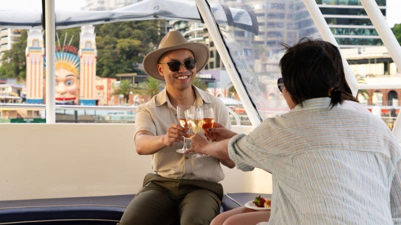 Pair toasts wine glasses aboard a Sydney Harbour catamaran at sunset, BYO cruise, city skyline illuminated in the background.