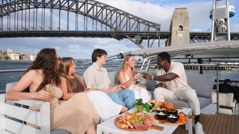 Friends enjoying gourmet food and drinks on a Sydney Harbour Golden Glow Sunset Cruise with iconic Sydney Bridge skyline views.