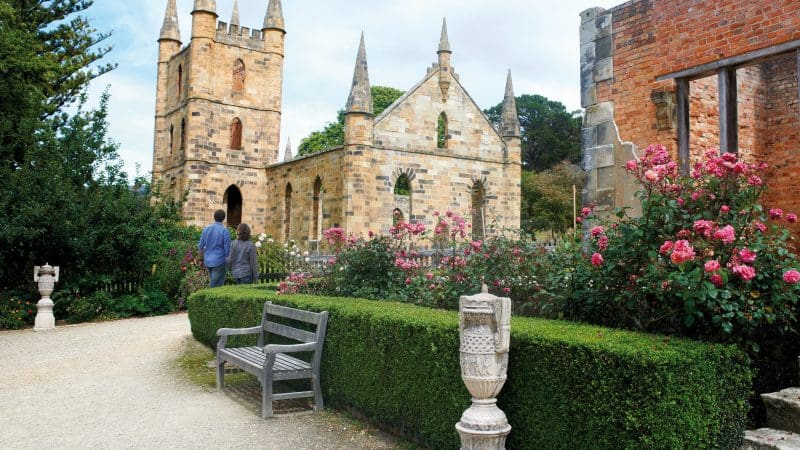 Discover the iconic sandstone church ruins with spires, lush gardens, and a rustic wooden bench on the 6 Day Tasmania Explorer Tour.