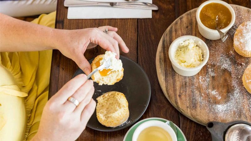 A person enjoys cream and jam on a scone during a CSHD Tour at a wooden table in Cairns City.