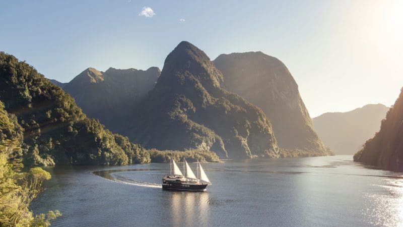 A serene sailboat glides on a tranquil lake, framed by majestic mountains and a bright, sunlit blue sky for a peaceful escape.