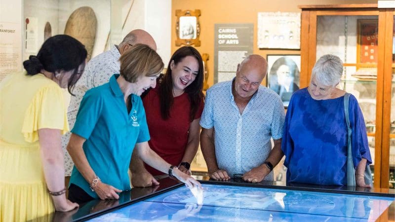 Six adults gather around an interactive touchscreen display during a CSHD Tour in a brightly lit Cairns City museum.