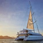 A white catamaran sails at sunset on calm waters during a CSDC Dinner Cruise, with mountains in the background.