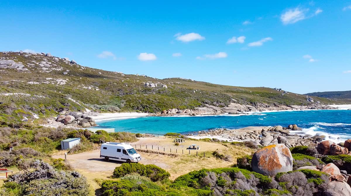 A campsite set on the beachfront bay. The bay has bright blue water and green hills rolling behind. A campervan is parked in the campsite.