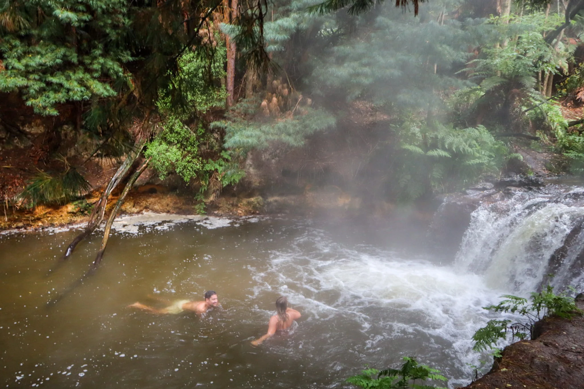 two people swimming in kerosene creek with steam coming from the water and waterfall