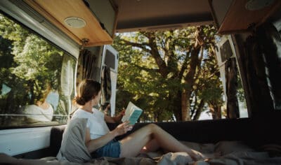 Girl sitting inside her Mighty campervan reading a book with the boot open and native trees behind them.