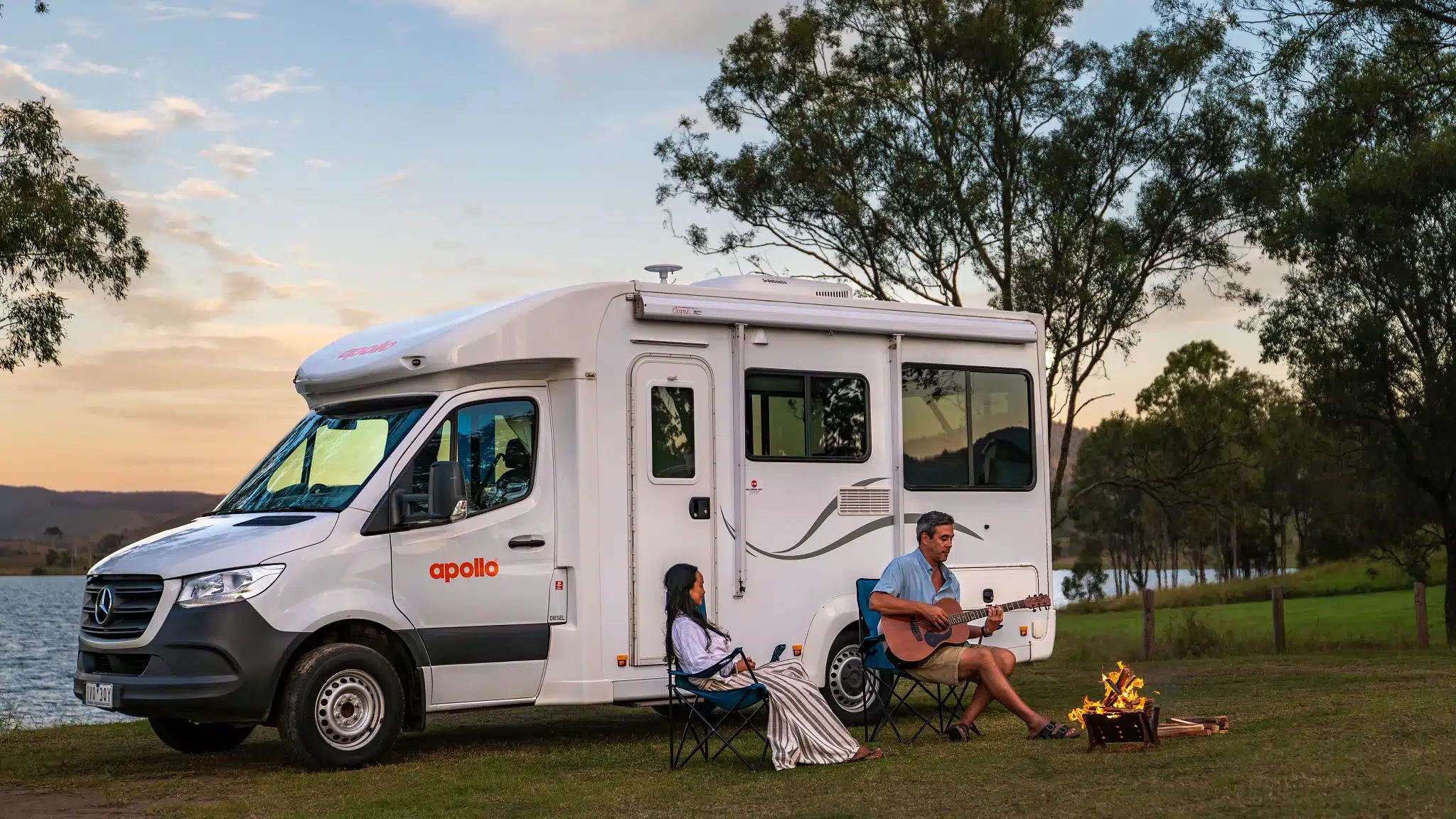 A RV Camper from Apollo Rentals is parked in front of a lake at sunset. A man and a woman sit in front of the RV while a campfire burns and the man plays guitar.