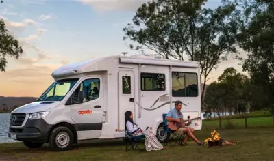 RV Motrohome from Apollo Rentals, parked by a lake with two people sitting under the awning playing a guitar.