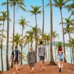 Four travellers stroll under tall palms towards the turquoise ocean on a sunny tropical beach in India and Sri Lanka.